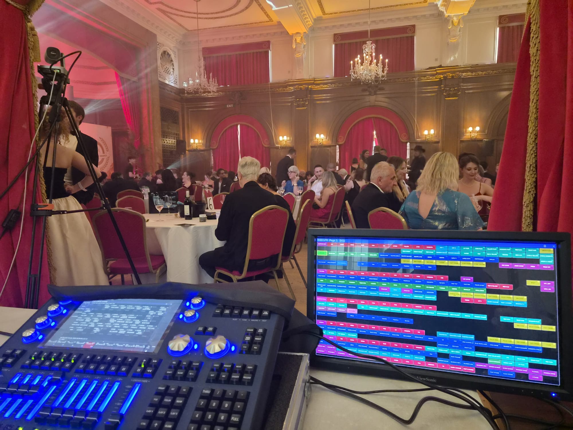 A lighting control desk and monitor set up at the back of a grand ballroom, with guests seated at tables in evening dress under chandeliers.